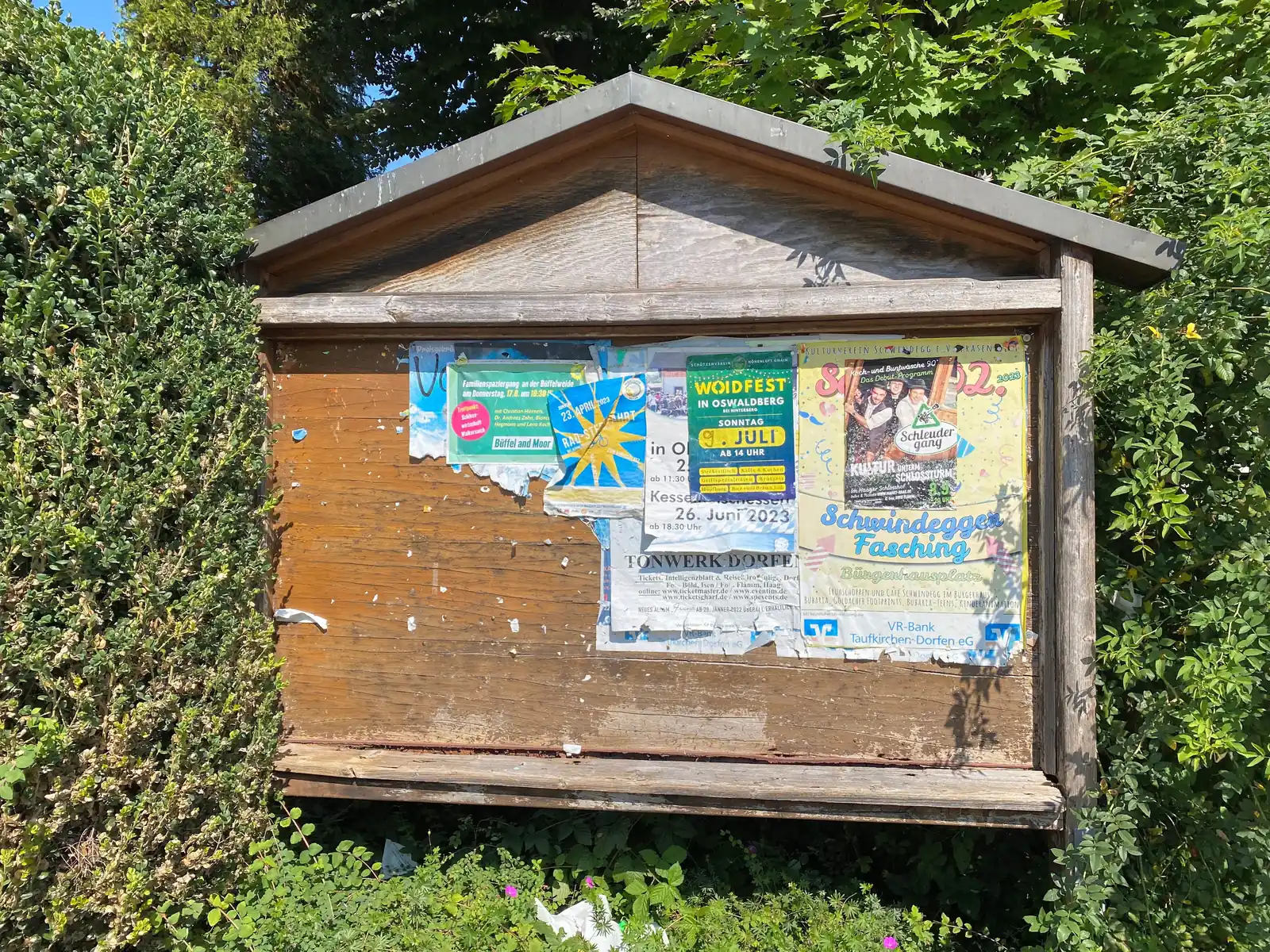 Hölzerner Anschlagkasten an einer Straße, mit Aushängen beklebt